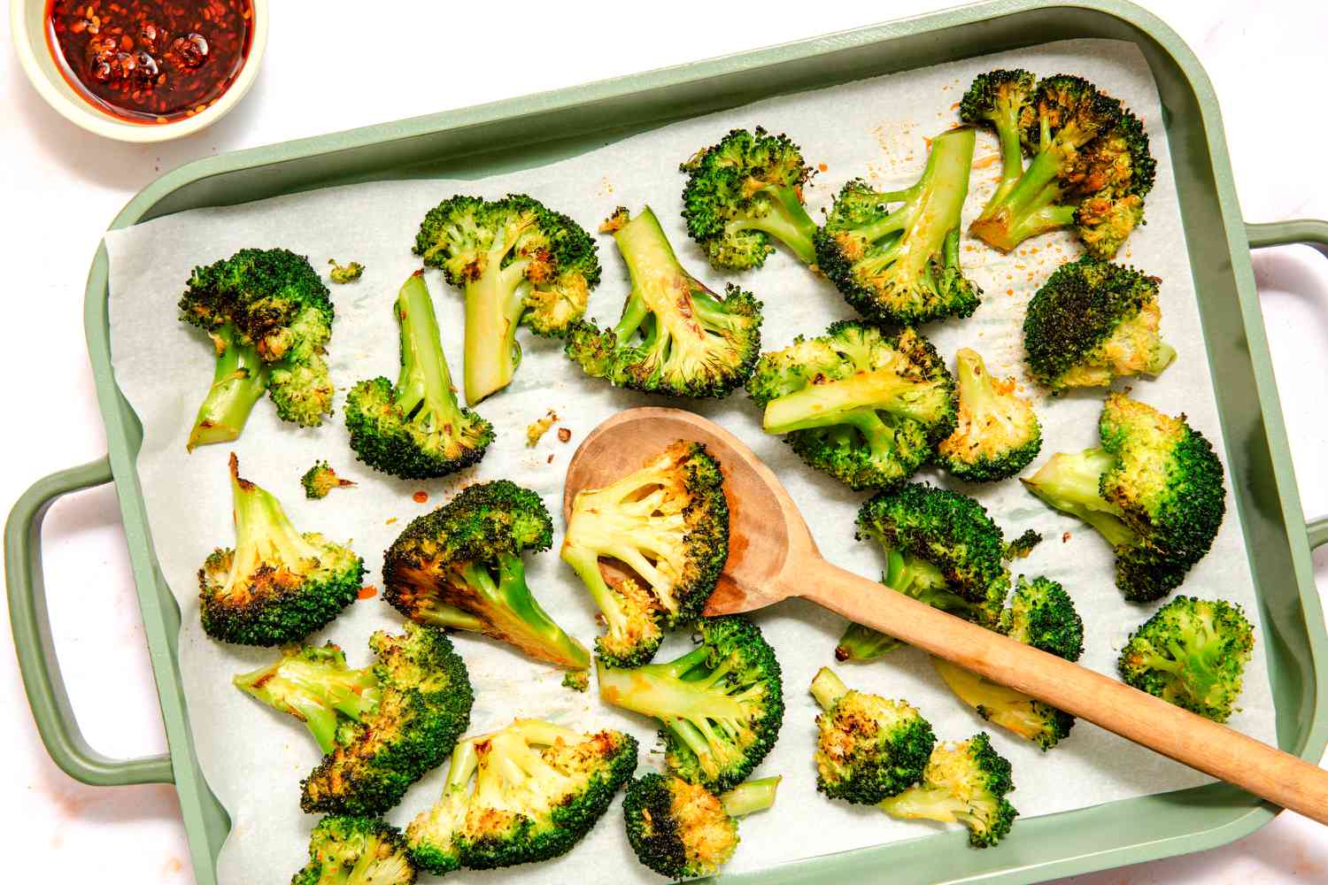 Vue de dessus d'une plaque de cuisson avec des fleurettes de brocoli recouvertes de chili croustillant sur du papier sulfurisé, avec une cuillère de service à côté d'un petit bol de chili croustillant