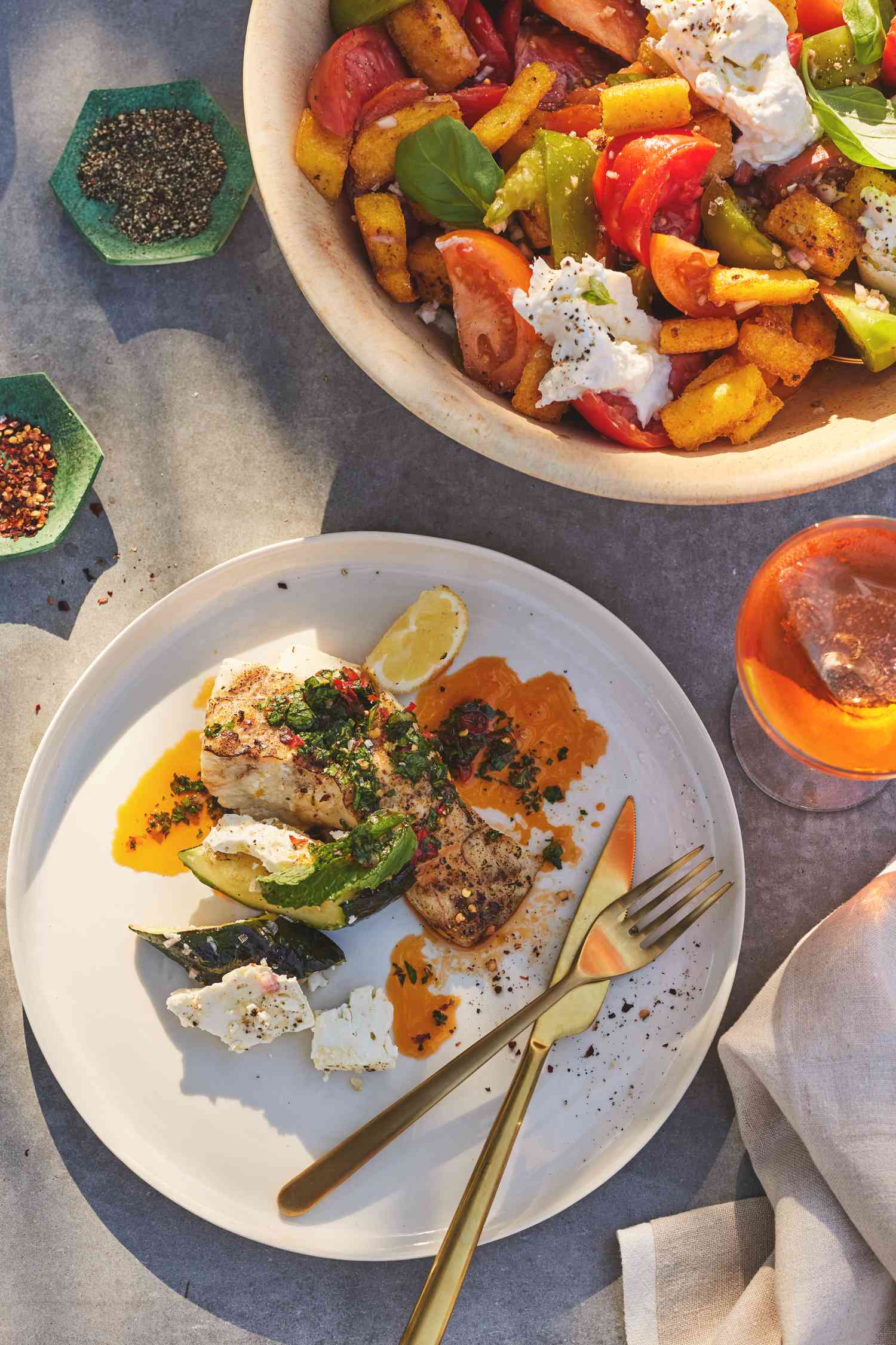 Portion sur une Assiette avec des Utensiles et un Bol de Panzanella de Polenta Frite avec Tomate, Basilic et Burrata entouré d'Épices et d'une Boisson sur une Table