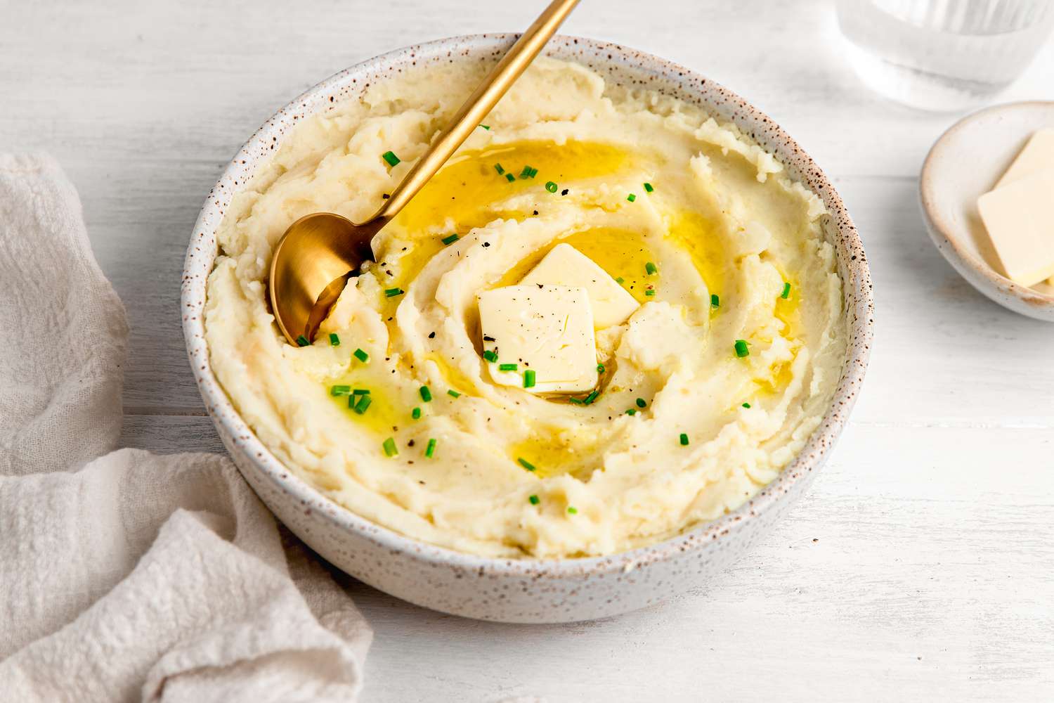 Vegan Mashed Potatoes in a Bowl Topped with Butter, Cracked Pepper, and Chives Next to a Saucer with More Butter