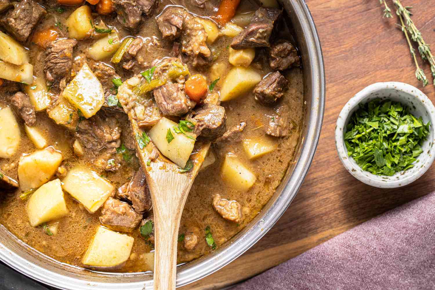 Pot de ragoût de bœuf facile avec une spatule en bois, un petit bol de persil, une branche de thym et un torchon de cuisine