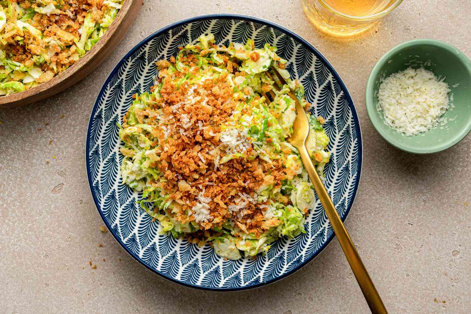 Salade César aux choux de Bruxelles avec des croûtons citronnés dans un bol avec une fourchette à côté d'un bol plus grand avec plus, un bol avec du parmesan râpé et un verre