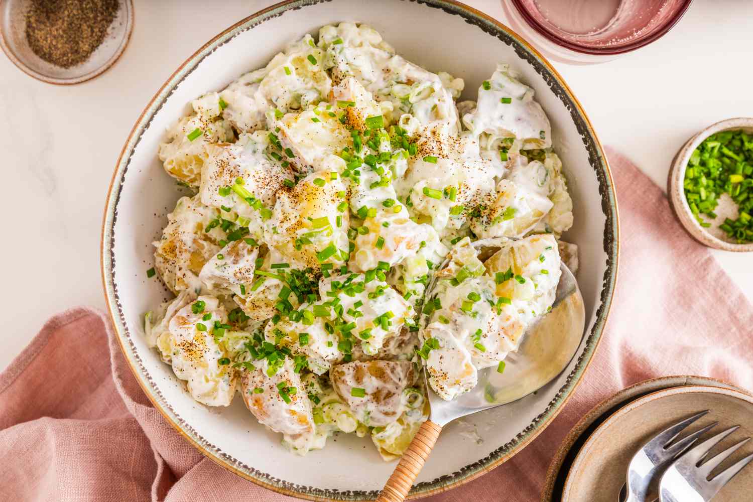 Bol de salade de pommes de terre à la crème aigre et à l'oignon avec une cuillère, entouré d'un bol d'assaisonnement, d'un verre d'eau, d'un bol de ciboulette, d'une pile d'assiettes avec des ustensiles, et d'une serviette de table rosée