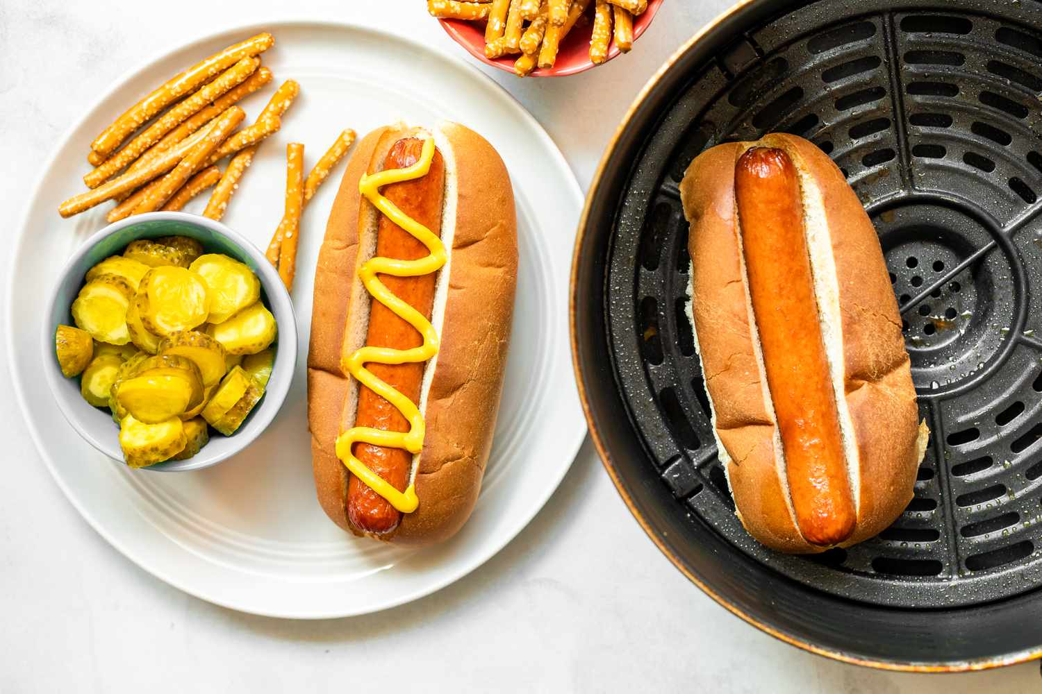 Une assiette de hot dog à la friteuse à air dans un pain garni de moutarde avec un accompagnement de cornichons et de bretzels.